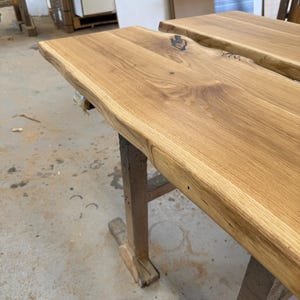 May include: A light brown wooden slab with a natural live edge and visible wood grain patterns. The surface features a dark knot detail and is supported by a wooden trestle base. The wood has a smooth, finished appearance with warm tones.
