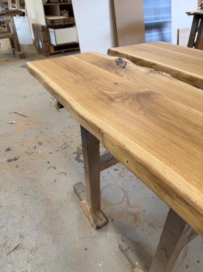 May include: A light brown wooden slab with a natural live edge and visible wood grain patterns. The surface features a dark knot detail and is supported by a wooden trestle base. The wood has a smooth, finished appearance with warm tones.