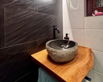 May include: A natural stone vessel sink with a rough, textured exterior and a smooth, polished interior basin. The sink is mounted on a thick, live-edge wooden slab countertop, paired with a matte black faucet and a light blue towel hanging below.