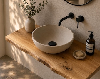 May include: A round, light-colored stone vessel sink sits on a natural live-edge wooden shelf. The sink features a matte black drain, paired with a matching wall-mounted matte black faucet and handle. A small vase with greenery and soap are present.