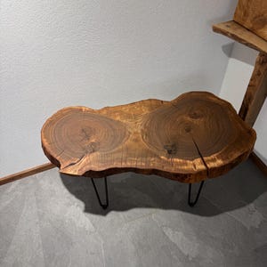 Könnte beinhalten: Ein Couchtisch mit lebendiger Kante aus einem einzigen Holzstück. Der Tisch hat eine natürliche, organische Form mit sichtbarer Holzmaserung und wird von schwarzen Haarnadelbeinen aus Metall getragen. Der Tisch ist etwa 102 cm lang.