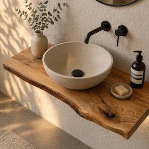 Puede incluir: Un moderno conjunto de lavabo de baño con un lavabo redondo de piedra beige claro sobre una estantería de madera natural. Un grifo negro y un dispensador de jabón complementan el diseño. Un pequeño jarrón con vegetación añade un toque natural.