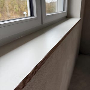 Könnte beinhalten: Nahaufnahme einer weißen Fensterbank mit Holzkante, unter einem weißen Fensterrahmen. Durch das Fenster sind Bäume und Himmel zu sehen. Die Wand ist hellbeige.