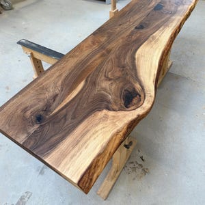 Pode incluir: Uma mesa de madeira com borda viva com uma forma natural e orgânica. A mesa é feita de madeira marrom escura com detalhes em marrom e bege mais claros. O grão da madeira é visível e a mesa é suportada por cavaletes de madeira.