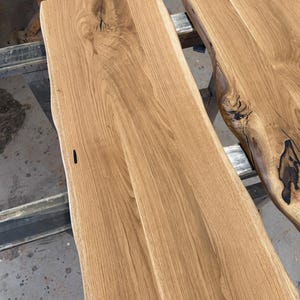 May include: Two rectangular light brown wooden boards with natural live edges. The wood grain is visible throughout the surface, featuring dark knots and organic imperfections that highlight the natural texture of the timber planks.
