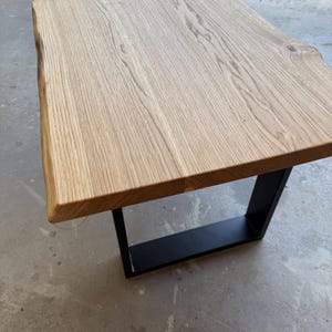 May include: A rectangular coffee table featuring a light-colored natural wood slab top with a live edge on one side. The table is supported by a sturdy, black powder-coated metal U-shaped base that provides a stable foundation for the wooden surface.