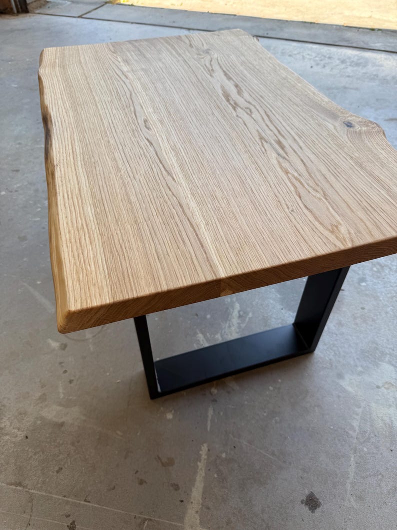 May include: A rectangular coffee table featuring a light-colored natural wood slab top with a live edge on one side. The table is supported by a sturdy, black powder-coated metal U-shaped base that provides a stable foundation for the wooden surface.