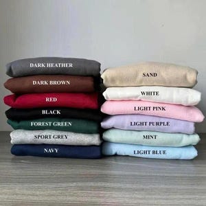 Pu&ograve; includere: Una pila di felpe piegate in vari colori: grigio scuro, marrone scuro, rosso, nero, verde foresta, grigio sportivo, blu navy, sabbia, bianco, rosa chiaro, lilla chiaro, menta e azzurro. Ogni felpa ha un'etichetta con il nome del colore.