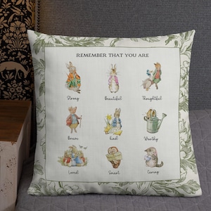 Puede incluir: Cojín cuadrado con un borde floral y un panel central con ilustraciones de acuarela de conejos. El panel incluye el texto "REMEMBER THAT YOU ARE" encima de las ilustraciones, con palabras como "Fuerte", "Hermoso" y "Amable" debajo.