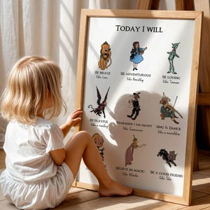 May include: A framed print with the text "TODAY I WILL" and illustrations of characters from The Wizard of Oz. The print is in a light wood frame and is propped up against a wall. A young child is sitting next to the print.