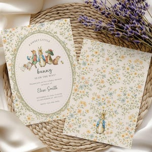May include: Baby shower invitations featuring a bunny theme. The front invitation has an oval frame with bunny illustrations and text. The back of the invitation has a floral pattern and a bunny illustration. The invitations are on a woven mat.