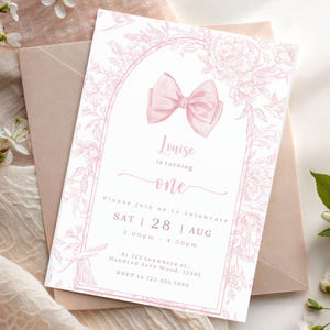 Puede incluir: Una tarjeta de invitación rosa con una ilustración de lazo y borde floral. El texto dice "Louise cumple un año" con detalles del evento: sábado 28 de agosto, 14:00-16:30. Se incluye información de RSVP. La tarjeta está sobre un sobre rosa.