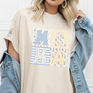 Puede incluir: Camiseta color crema con la palabra "MAMA" en letras grandes estampadas. Las letras están decoradas con diseños de conejitos y huevos de Pascua en azul, amarillo y blanco. La camiseta tiene mangas cortas y cuello redondo.