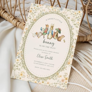 Puede incluir: Invitación a un baby shower con un diseño floral y el texto "A sweet little bunny is on the way!". La invitación presenta ilustraciones de conejos y otros animales en un marco ovalado. La invitación es de color crema con un borde floral.
