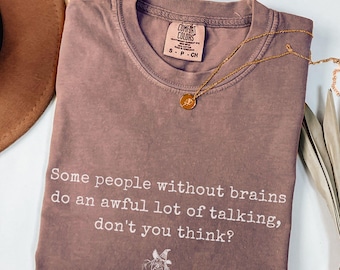 May include: A mauve Comfort Colors t-shirt with the text "Some people without brains do an awful lot of talking, don't you think?" and a small illustration. A gold necklace and a brown hat are also visible.