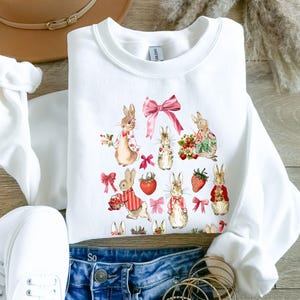 Op de afbeelding: Witte crewneck sweatshirt met een aquarelontwerp van konijntjes, aardbeien en roze strikken. De konijntjes zijn gekleed in verschillende outfits. De sweatshirt is gemaakt van een zacht materiaal, ideaal voor de lente of Pasen.