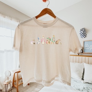 May include: Beige short-sleeved t-shirt featuring a watercolor illustration of several animal characters. The design includes a rabbit, duck, and other figures. The shirt is hanging on a wooden hanger.