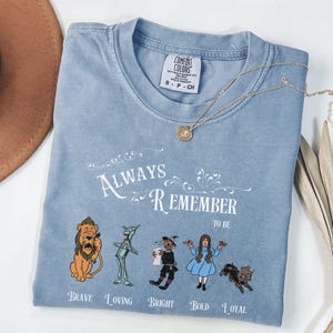 May include: A light blue Comfort Colors t-shirt with the text "Always Remember to Be" and illustrations of characters from The Wizard of Oz. A gold necklace with a pendant is draped across the shirt.