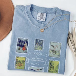 May include: A pale blue Comfort Colors t-shirt with a design of vintage postage stamps featuring illustrations and the text "WE'RE NOT IN KANSAS ANYMORE." A gold necklace with a pendant is on the shirt.