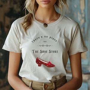 Op de afbeelding: Een wit T-shirt met de tekst "There's no place like The Shoe Store" en een rode glinsterende schoen met een strik.