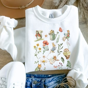 Peut inclure: Sweat-shirt blanc avec un imprimé coloré d'animaux de la forêt, de fleurs et d'un arrosoir. L'imprimé comprend un écureuil, un lapin, une souris, un hibou et un papillon.