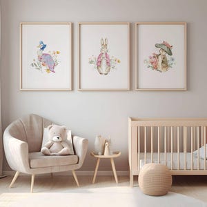 Può includere: Tre stampe ad acquerello incorniciate con personaggi di libri per bambini. Le stampe raffigurano un'anatra, un coniglio e un topo, ciascuno circondato da fiori. Le stampe sono in cornici di legno chiaro, esposte sopra una sedia beige, un tavolino e una culla.