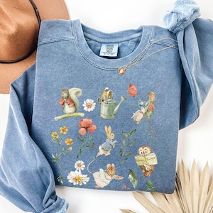 Peut inclure: Un sweat-shirt bleu avec un imprimé d'inspiration vintage représentant un écureuil, un lapin, une souris, un oiseau, un arrosoir et des fleurs. L'impression est dans des tons de brun, vert, jaune et rouge.