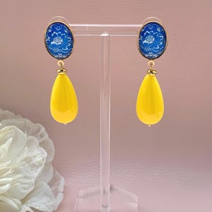 Könnte beinhalten: Ohrringe zum Hängen mit ovalen, blau-weiß geblümten Oberteilen und leuchtend gelben, tropfenförmigen Anhängern. Die Ohrringe werden auf einem transparenten Acrylständer präsentiert. Die Ohrringe haben goldfarbene Akzente.