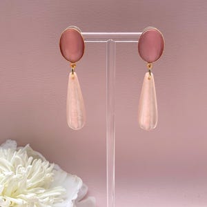 Könnte beinhalten: Ein Paar Ohrhänger mit ovalen, altrosa Cabochons in goldfarbenen Metallfassungen. An jedem ovalen Oberteil hängt ein langer, tropfenförmiger Anhänger in einem hellen, schimmernden, perlmuttartigen Hellrosa mit glänzender Oberfläche.