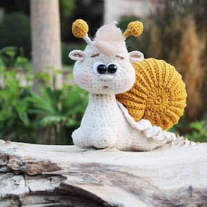 Puede incluir: Un juguete de ganchillo en forma de caracol con un cuerpo blanco y una concha amarilla. El caracol tiene grandes ojos negros y una nariz rosa.