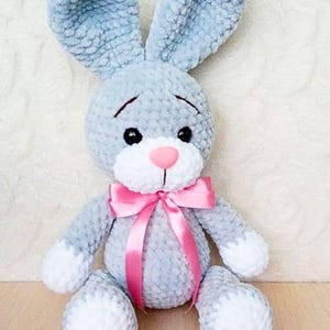 May include: A gray crocheted bunny rabbit plush toy with white feet and a pink satin bow. The bunny has black eyes and a pink nose.