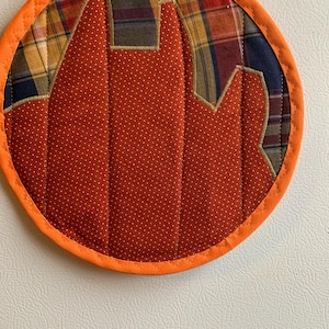 May include: A round orange pot holder with a quilted design. The centre of the pot holder is a brown fabric with white polka dots. The outer edge of the pot holder is a plaid fabric in shades of brown, yellow, and blue.