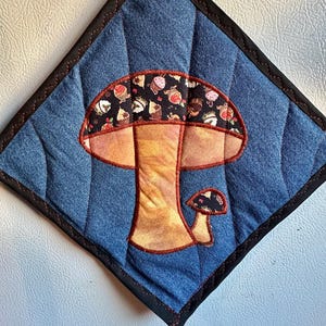 Puede incluir: Un salvamanteles acolchado de mezclilla con un diseño de seta. La seta tiene un tallo marrón y un sombrero decorado con cupcakes sobre un fondo negro. El salvamanteles cuadrado tiene un borde negro y costuras rojas.