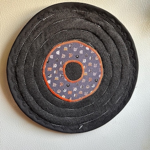 Può includere: Un sottobicchiere circolare in tessuto con un anello esterno in denim grigio scuro e cuciture concentriche bianche. Il centro è una toppa in tessuto viola chiaro con un motivo di piccole teste di gatto, bordata da una cornice arancione.