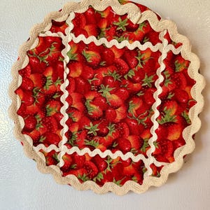 Peut inclure: Un dessous de verre rond en tissu rouge orné d'un motif de fraises. Le bord est festonné et orné d'une bordure crème. Le centre présente un motif répété de fraises mûres avec des feuilles vertes.