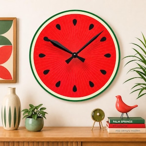 May include: A round wall clock featuring a vibrant red watermelon slice design with black seeds, a thin white rind, and a dark green outer border. The clock face has black hour and minute hands radiating from the center, set against a red background.