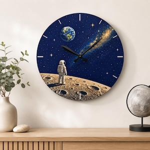 May include: A round wall clock featuring a space-themed graphic of an astronaut standing on the cratered surface of the moon, looking out at a dark blue starry sky with a visible Earth and a glowing golden nebula, complete with black clock hands.