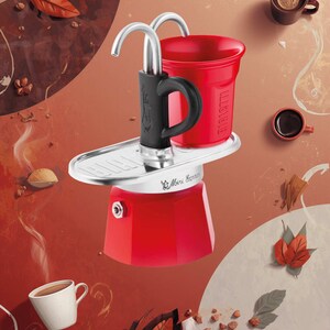 Może przedstawiać: Czerwony ekspres do kawy Mini Express z czarnym uchwytem i srebrnymi akcentami. Obraz zawiera filiżanki kawy, ziarna kawy i jesienne liście na czerwonobrązowym tle. Słowo "BIALETTI" jest widoczne na czerwonym kubku.