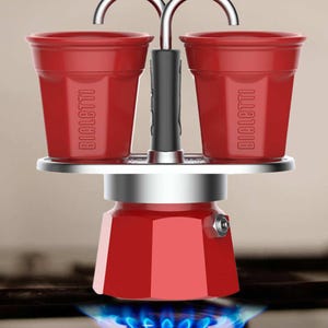 Może przedstawiać: Czerwony ekspres do kawy na kuchenkę z dwoma filiżankami, z logo "BIALETTI". Ekspres do kawy jest podgrzewany przez niebieski płomień gazu. Filiżanki są przymocowane do g&oacute;ry ekspresu do kawy.