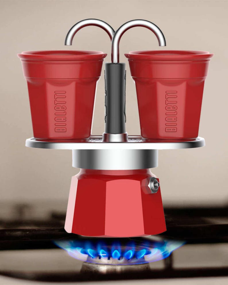Może przedstawiać: Czerwony ekspres do kawy na kuchenkę z dwoma filiżankami, z logo "BIALETTI". Ekspres do kawy jest podgrzewany przez niebieski płomień gazu. Filiżanki są przymocowane do g&oacute;ry ekspresu do kawy.