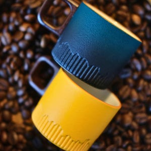 Peut inclure: Deux tasses en céramique empilées avec des anses en bois. L'une est bleu marine, l'autre jaune. Les deux tasses présentent un motif texturé et strié près de la base. Les tasses sont posées sur un fond de grains de café.