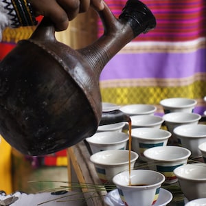 Op de afbeelding: Een persoon schenkt koffie uit een traditionele kleipot, genaamd jebena, in kleine witte kopjes. De kopjes zijn gerangschikt op een tafel met een bruin oppervlak. De achtergrond heeft een kleurrijk gestreept patroon.