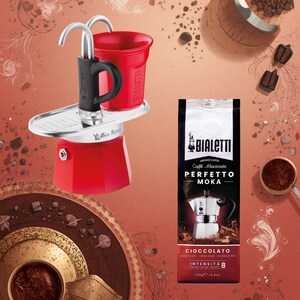 Może przedstawiać: Czerwony ekspres do kawy Bialetti z czarnym uchwytem i czerwonym kubkiem. Obok ekspresu znajduje się paczka kawy mielonej Bialetti z napisem "Perfetto Moka Cioccolato". Na zdjęciu znajduje się r&oacute;wnież czekolada i ziarna kawy.