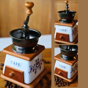 Puede incluir: Un molinillo de café manual vintage con un cuenco de metal oscuro y un mango de madera. La base de cerámica blanca presenta un cajón y la palabra "CAFE". Patrones florales y granos de café rodean el molinillo.