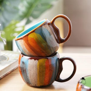 Puede incluir: Dos tazas de cerámica coloridas apiladas una encima de la otra. Las tazas tienen un esmalte marrón con rayas coloridas de azul, amarillo, naranja y verde. Las tazas tienen una forma redondeada y un asa.