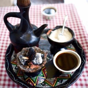 Puede incluir: Un conjunto tradicional de ceremonia de café etíope en una bandeja. Incluye una cafetera negra, una taza pequeña de café, un cuenco de azúcar y un pequeño quemador de incienso con carbón encendido. La bandeja tiene un diseño geométrico colorido.
