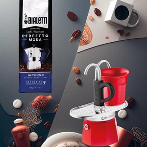 Może przedstawiać: Zdjęcie produktu przedstawiające czerwono-srebrny ekspres do kawy, paczkę kawy mielonej i czerwony kubek. Na opakowaniu kawy widnieje napis "PERFETTO MOKA" i "INTENSO". Ziarna kawy i białe cukierki są rozrzucone wok&oacute;ł przedmiot&oacute;w.