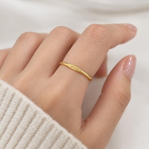 May include: A thin gold-toned ring featuring a small, flat, horizontal bar section at the centre. The bar is engraved with the name Lucy in a delicate, black cursive script. The band is smooth and polished, maintaining a consistent, slender profile.