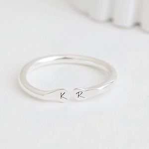 May include: A silver-toned open ring featuring a smooth, polished band with two rounded ends. The left end is engraved with the letter K, and the right end is engraved with the letter R in a simple, dark, sans-serif font style.