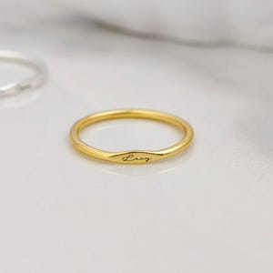 Op de afbeelding: Een goudkleurige ring met een dunne band en een klein, plat, ovaalvormig middengedeelte gegraveerd met de naam Lucy in cursief schrift, vergezeld van een tweede, effen zilverkleurige ring met een dunne, gladde, ronde band.